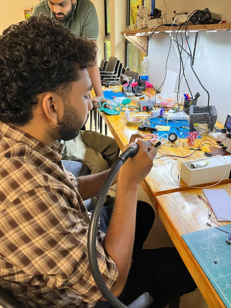 Muhammed Basheer at the workbench, working on a small device with a rotary tool, surrounded by wires and tools