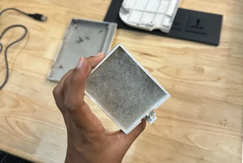 A hand holding the ventilator's square intake filter, its surface matted grey with dust
