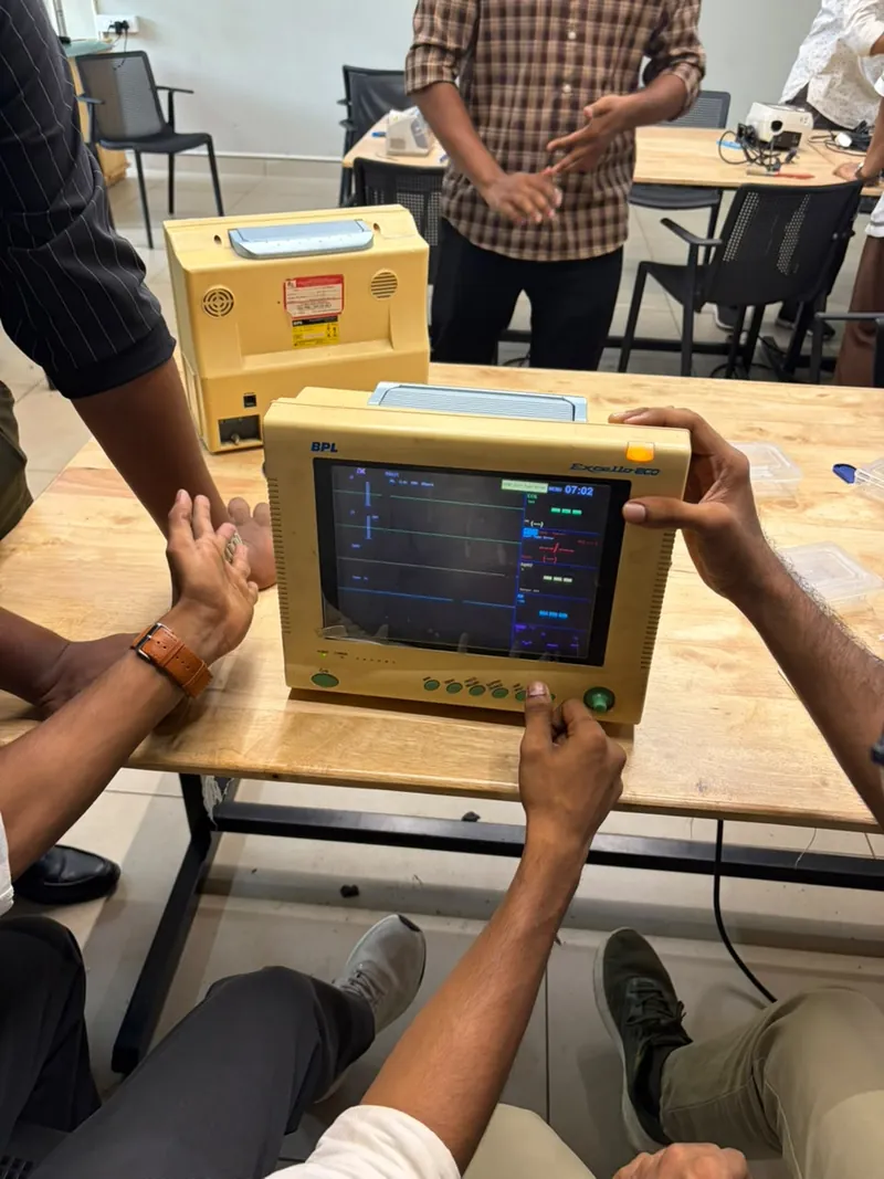 A yellow BPL Excello patient monitor switched on and showing traces, with hands around it pressing its buttons