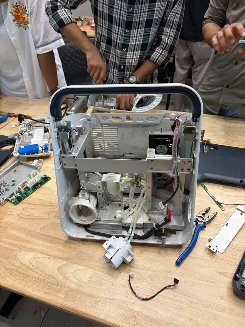 A Mindray SV300 ventilator with its casing removed, showing the metal frame, a fan, tubing and wiring, with tools around it on the table