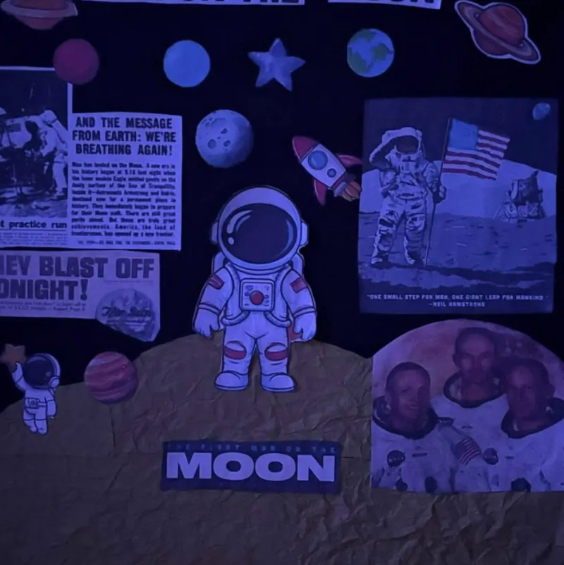 A display wall about the Moon landing with newspaper clippings, an astronaut cut-out standing on a paper Moon, and a photo of the Apollo 11 crew